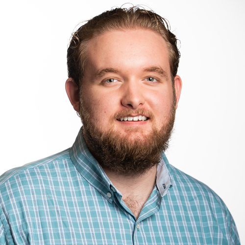 Portrait of Nic Jones smiling, with a beard, wearing a light blue checkered shirt.