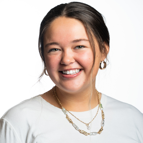 Headshot of Kelsey Robinett, a smiling young woman with long dark hair, wearing a white shirt and layered necklaces.