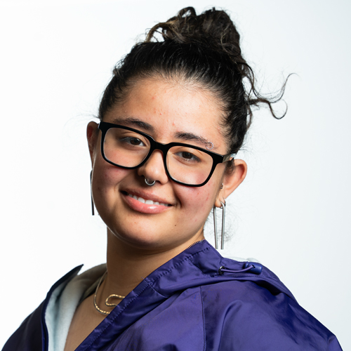 Portrait of AJ Ware, a young woman with glasses and a bun hairstyle, smiling and wearing a purple jacket.